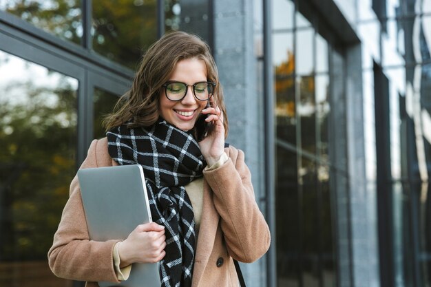 Wizerunek pięknej młodej kobiety na zewnątrz spaceru ulicą trzymając laptopa rozmawia przez telefon komórkowy.