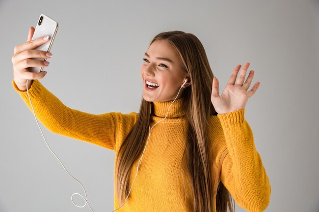 Wizerunek pięknej szczęśliwej młodej kobiety na białym tle nad szarą ścianą przy użyciu telefonu komórkowego mówi macha.