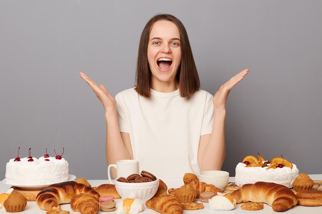 Wizerunek podekscytowanej radosnej wesołej kobiety z brązowymi włosami siedzącej przy stole odizolowanej na szarym tle z mnóstwem pysznych deserów krzyczących ze szczęścia patrząc na kamerę