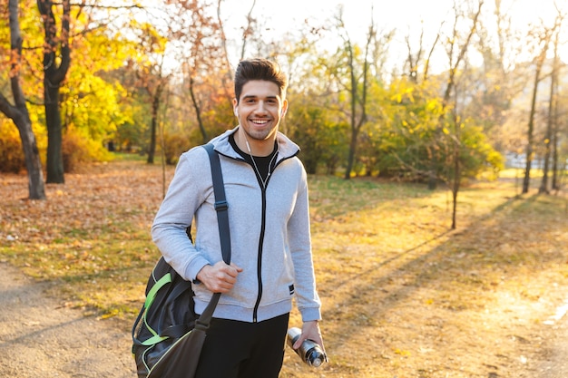 Wizerunek przystojnego młodego sportowca na świeżym powietrzu w parku, słuchanie muzyki ze słuchawkami, chodzenie z torbą.