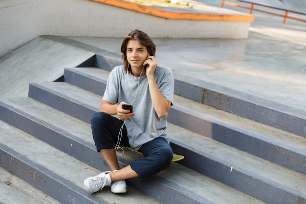 Wizerunek Przystojny Atrakcyjny Młody Skater Siedzieć W Parku, Słuchając Muzyki Przez Słuchawki Przy Użyciu Telefonu.