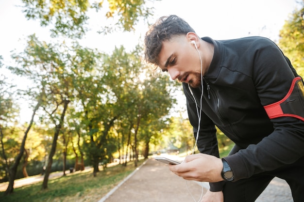 Wizerunek przystojny młody sport fitness biegacz mężczyzna na zewnątrz w parku słuchanie muzyki ze słuchawkami za pomocą telefonu komórkowego.