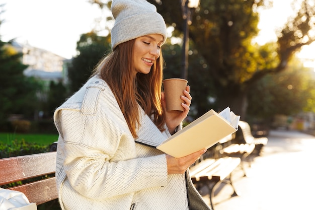 Wizerunek rudowłosej kobiety siedzącej na ławce na zewnątrz trzymając książkę, pijąc kawę.