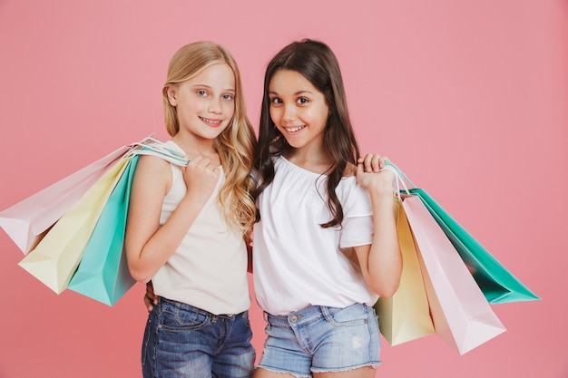 Wizerunek ślicznej brunetki i blondynki w wieku 8-10 lat w zwykłym ubraniu, uśmiecha się do kamery i trzyma kolorowe torby na zakupy z zakupami, odizolowane na różowym tle