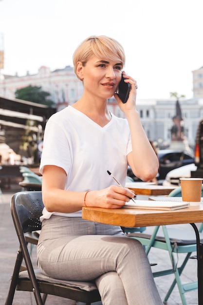 Wizerunek stylowej dojrzałej kobiety noszącej podstawowe ubrania, zapisującej w notatniku i rozmawiającej przez telefon komórkowy, siedząc w letniej kawiarni na świeżym powietrzu