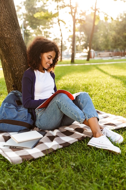 Wizerunek szczęśliwy afrykańskiej młodej kobiety siedzącej na świeżym powietrzu w parku, pisanie notatek w zeszycie, słuchanie muzyki przez słuchawki.