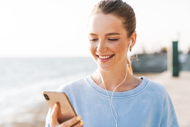 Wizerunek wesołej pozytywnej młodej kobiety fitness na zewnątrz na plaży przy użyciu telefonu komórkowego słuchania muzyki ze słuchawkami.