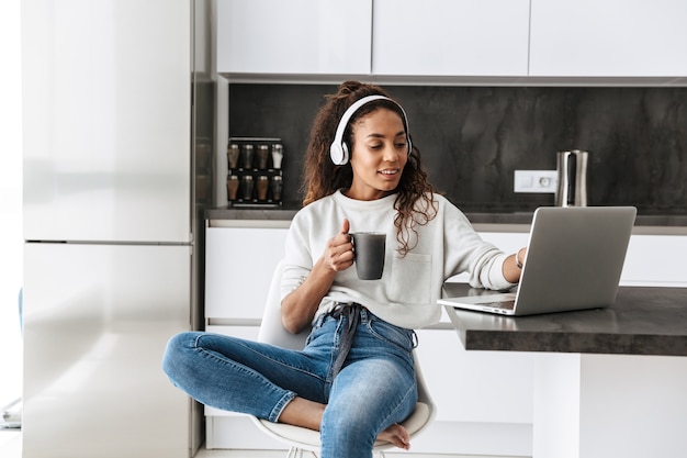 Wizerunek Współczesnej African American Girl Noszenia Słuchawek Za Pomocą Laptopa, Siedząc W Jasnej Kuchni