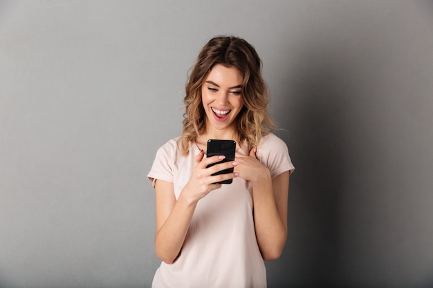 Wizerunek Zadowolona kobieta w koszulki writing wiadomości na smartphone nad popielatym