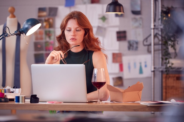 Właścicielka firmy modowej z lampką wina pracująca do późna na laptopie w studio