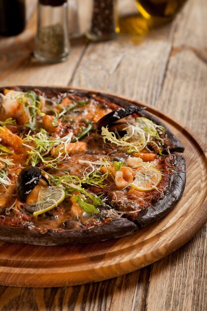 Włoska pizza "Di Mare" z czarnym ciastem i owocami morza na drewnianym stole. Pojemniki z przyprawami i butelką oliwy z oliwek. Na wierzchu połóż krewetki, cytrynę, rukolę, sałatę i otwartą skorupkę z małżami.