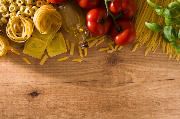 Włoski makaron i składniki. Ravioli, makaron penne, spaghetti, tortellini, pomidory i bazylia na drewnianym stole