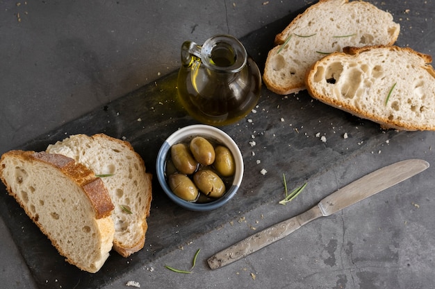 Włoski Pokrojony Chleb Ciabatta Na Desce Do Krojenia Z Ziołami, Oliwą Extra Virgin I Marynowanymi Oliwkami Na Ciemnym Tle Grunge