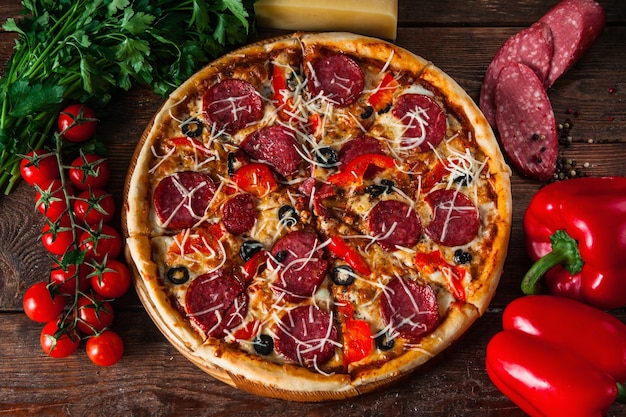 Włoskie jedzenie. Gorąca pizza na stole ze składnikami. Pieprz, kiełbasa, wiśnie, parmezan i zielona pietruszka.