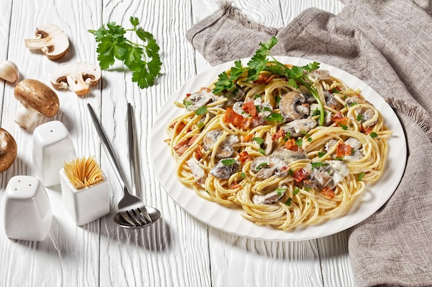 Włoskie spaghetti z kremowym sosem śmietanowo-parmezanowym z chrupiącym boczkiem i pietruszką podane na białym talerzu i sztućcach na białym drewnianym tle, widok z góry, zbliżenie