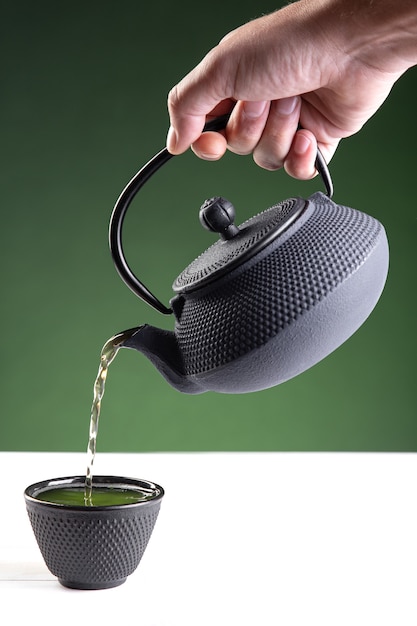 Woda wlewa się do ciemnej filiżanki zielonej herbaty na zielonym tle z kopią miejsca.Format pionowy.