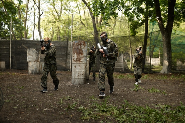 Wojownicy w kamuflażu i maskach grający w paintball. Sporty ekstremalne z bronią pneumatyczną i kulami lub markerami malarskimi, wojskowa gra zespołowa w plenerze, taktyka walki