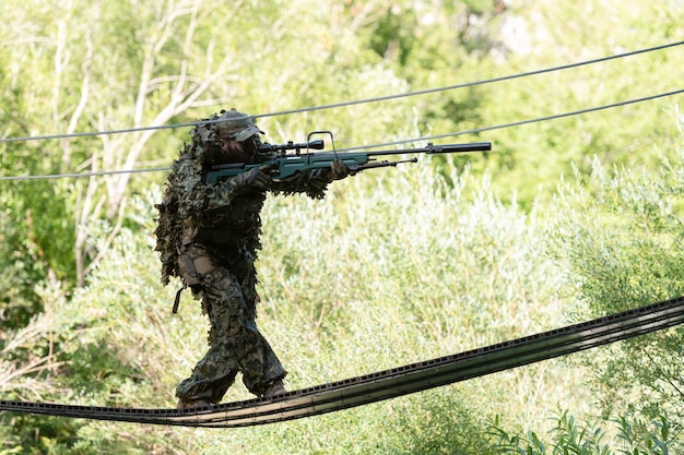 Wojskowy lub gracz airsoftowy w kostiumie kamuflażowym skrada się po moście linowym i celuje z karabinu snajperskiego w bok lub do celu.