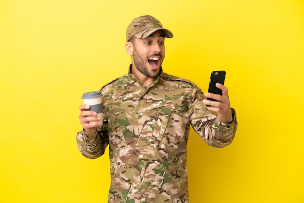 Wojskowy odizolowany na żółtym tle trzymający kawę na wynos i telefon komórkowy