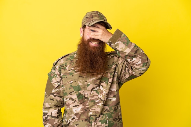 Zdjęcie wojskowy rudy mężczyzna na białym tle na żółtym tle zakrywający oczy rękami i uśmiechnięty
