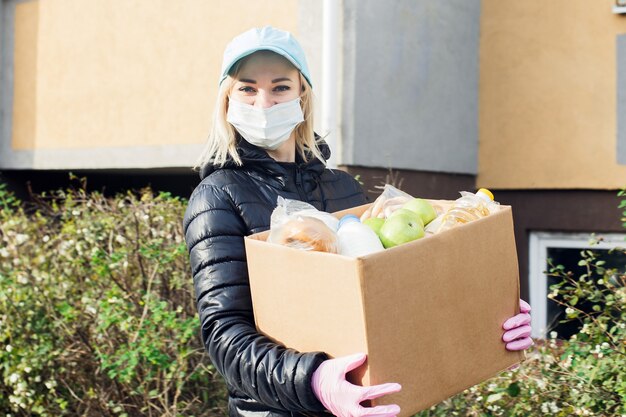 Wolontariuszka w masce ochronnej idzie ulicą z pudełkiem zakupów i darowizn.