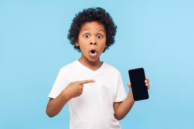 Wow niesamowita aplikacja mobilna. Zdziwiony podekscytowany ładny mały chłopiec wskazujący na telefon komórkowy i patrzący na aparat z zszokowaną twarzą, dziecko zaskoczony telefonem, usługa online. kryty studio strzał na niebieskim tle