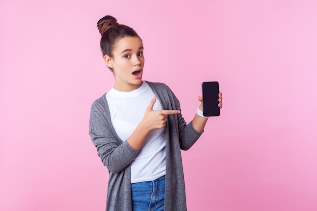 Wow, spójrz na aplikację mobilną. Portret zdziwionej nastoletniej brunetki z kok fryzurą w zwykłych ubraniach, wskazując na telefon, zdumiony zszokowany gadżetem. studio strzał na białym tle na różowym tle