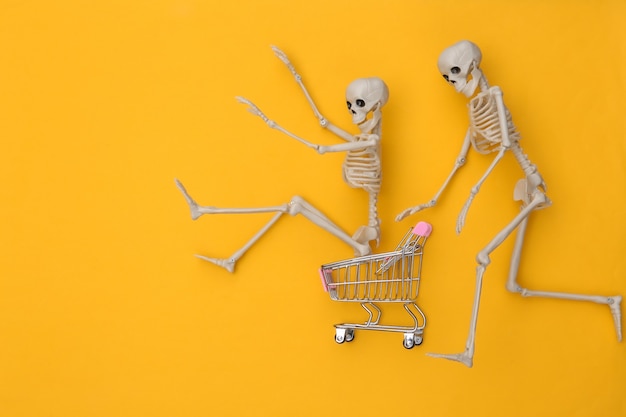 Zdjęcie wózek na zakupy z śmiesznymi szkieletami na żółtym tle. motyw halloween. widok z góry