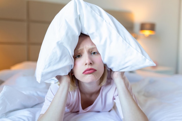 Wściekła kobieta cierpiąca z powodu hałasu sąsiada leżąca w nocy na łóżku zakrywająca uszy poduszką z powodu dźwięku