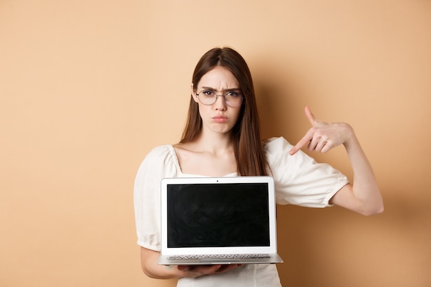 Wściekła, marszcząca brwi kobieta w okularach wskazująca na ekran laptopa, narzekająca i wyglądająca na rozczarowaną, stojąca na beżowym tle.