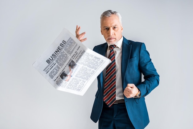Zdjęcie wściekły dojrzały mężczyzna w formalnej odzieży rzuca gazetę biznesową, stojąc odizolowany na szarym