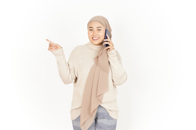 Wskazując na bok i rozmawiając przez telefon pięknej azjatyckiej kobiety noszącej hidżab na białym tle