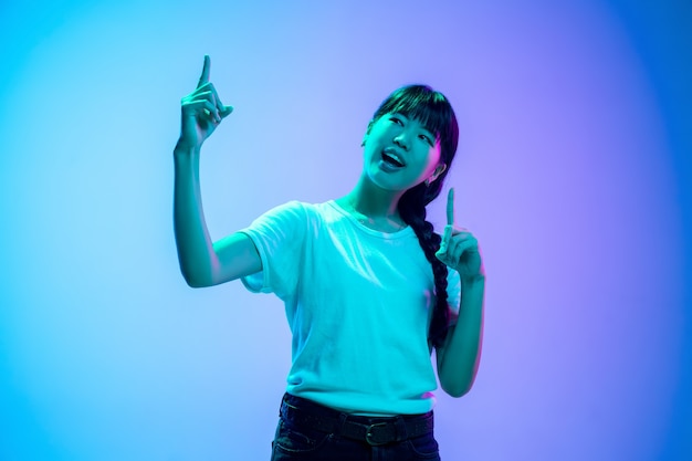 Wskazując w górę. Portret młodej kobiety azjatyckiej na gradientowym niebiesko-fioletowym tle studio w świetle neonowym. Pojęcie młodości, ludzkie emocje, wyraz twarzy, sprzedaż, reklama. Piękna brunetka modelka.