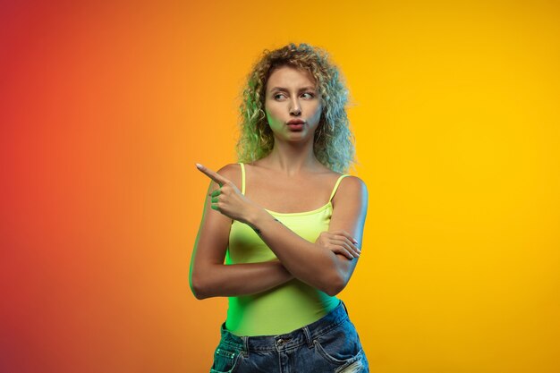 Wskazywanie. Portret młodej kobiety rasy kaukaskiej na tle gradientu studio w neon. Piękna modelka kręcone w stylu casual. Pojęcie ludzkich emocji, wyraz twarzy, młodość, sprzedaż, reklama.