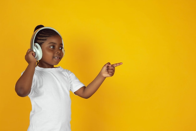 Wskazywanie, słuchanie muzyki. Portret małej afro-amerykańskiej dziewczyny na żółtym tle studio. Wesoły dzieciak. Pojęcie ludzkich emocji, wyraz twarzy, sprzedaż, reklama. Miejsce. Wyglądać słodko.