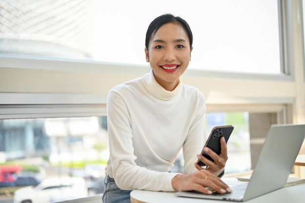 Wspaniała azjatycka kobieta siedzi w kawiarni ze swoim smartfonem i laptopem uśmiechając się i patrząc na kamerę