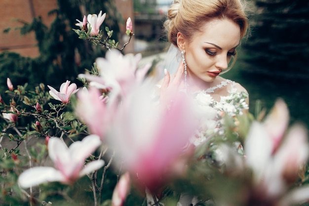 Zdjęcie wspaniała blondynka panna młoda pozuje w pobliżu kwiatów magnolii nowożeńcy portret panny młodej piękna kobieta w białej sukni pozowanie w pobliżu kwiecistego krzewu na zewnątrz
