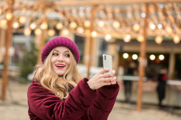 Wspaniała blondynka ubrana w ciepłe ubrania robi selfie na tle girlandy w Kijowie