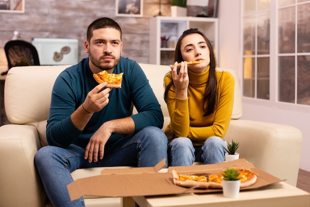 Wspaniała młoda para je pizzę podczas oglądania telewizji w salonie siedząc na kanapie