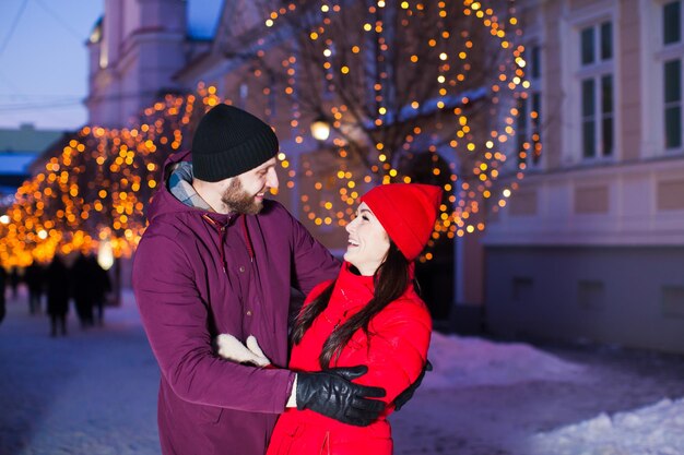 Wspaniała młoda żonata rodzina przytula się na świeżym powietrzu zimą udekorowane miasto w zimną noc, ubrana ciepło w nakrycia głowy, rękawiczki, płaszcze, patrząc na siebie z pasją