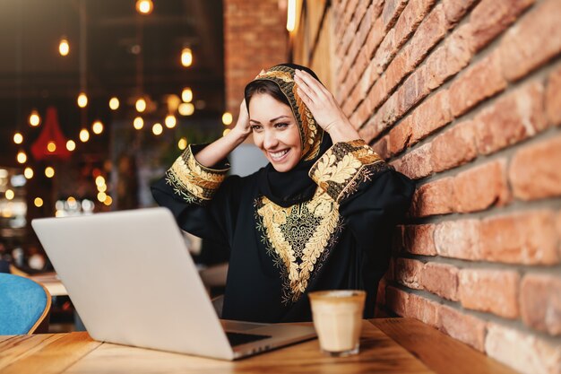 Wspaniała Muzułmańska Kobieta Z Zębatym Uśmiechem Ubierał W Tradycyjnej Odzieży Używać Laptop Do Zakupów On-line Podczas Gdy Siedzący W Bufecie. Kawa Na Biurku.