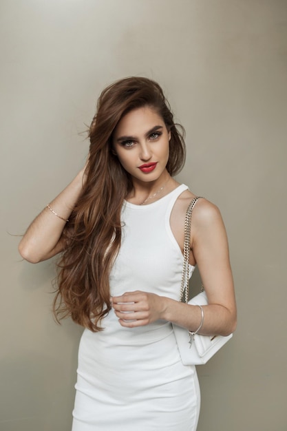 Wspaniała, piękna młoda kobieta w modnej, eleganckiej białej sukience z torbą stoi przy beżowej ścianie i prostuje włosy