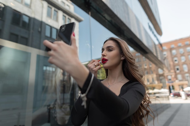 Zdjęcie wspaniała piękna młoda moda szykowna dziewczyna modelka maluje usta czerwoną szminką i patrzy na smartfon w mieście