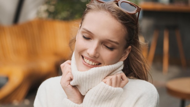 Wspaniała uśmiechnięta dziewczyna ubrana w przytulny sweter z białym kołnierzykiem, która wygląda uroczo, ciesząc się ciepłą pogodą w ulicznej kawiarni