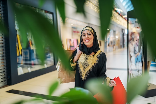 Wspaniała uśmiechnięta młoda Arabka w tradycyjnej odzieży stojącej w centrum handlowym z torby na zakupy w ręce