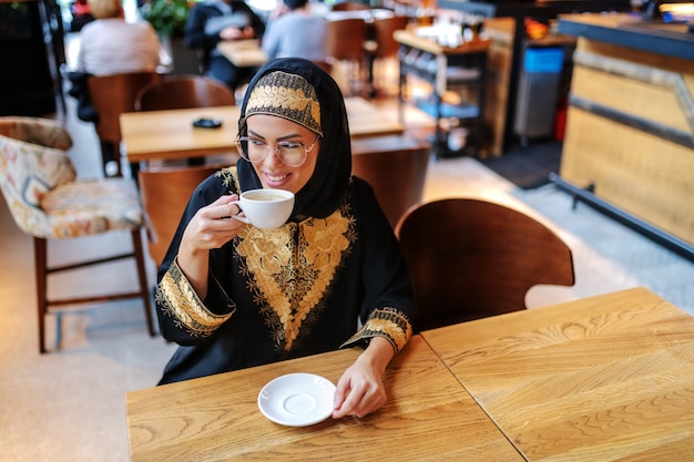 Wspaniała uśmiechnięta pozytywna Arabka w tradycyjnej odzieży siedząca samotnie w kawiarni
