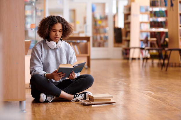 Współczesna dziewczyna w obcisłych dżinsach i szarej bluzie czytająca książkę siedząc na podłodze w bibliotece uczelni