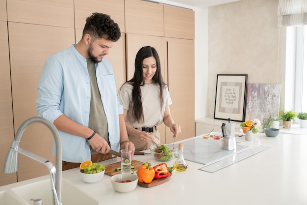 Współczesna młoda para w stroju codziennym, stojąca przy kuchennym stole i gotująca śniadanie składające się z sałatki warzywnej z ogórkami
