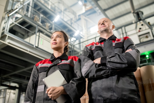 Współcześni inżynierowie płci męskiej i żeńskiej w odzieży roboczej stojącej przed kamerą na tle ogromnej konstrukcji technicznej w dużym magazynie