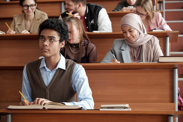 Współcześni międzykulturowi studenci uniwersyteccy robiący notatki siedząc przy długich drewnianych ławkach w sali wykładowej i słuchający profesora na lekcji